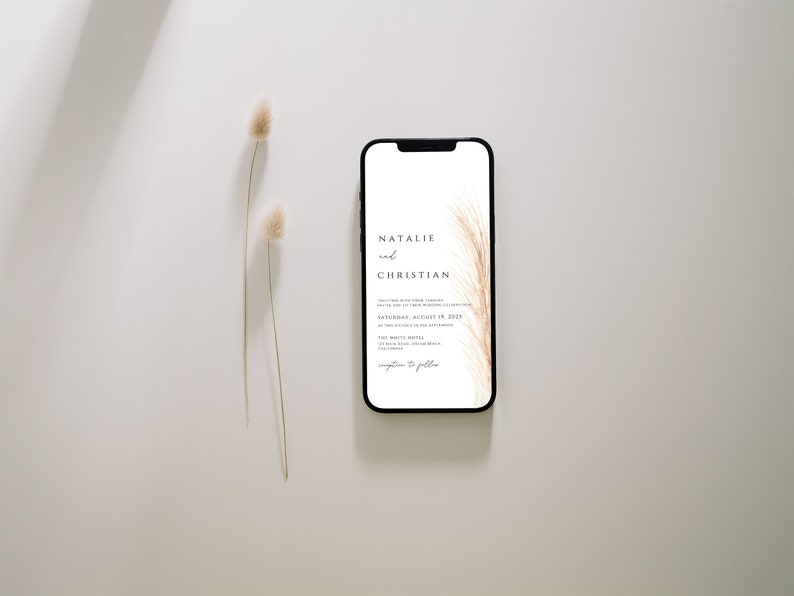 May include: A smartphone displays a wedding invitation with the names "Natalie and Christian" in elegant black script. The invitation features a soft, feathery design in shades of beige and brown. Two dried flower stems are positioned to the left of the phone.
