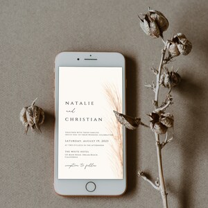 May include: A smartphone displays a wedding invitation with the names "Natalie and Christian" and event details. The invitation features a minimalist design with a beige background and elegant typography. Dried floral elements frame the phone.
