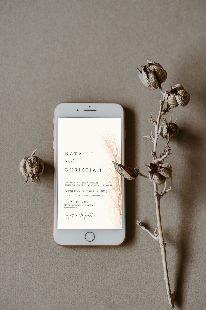 May include: A smartphone displays a wedding invitation with the names "Natalie and Christian" and event details. The invitation features a minimalist design with a beige background and elegant typography. Dried floral elements frame the phone.