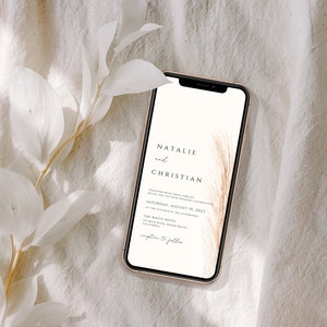 May include: A smartphone displays a wedding invitation with the names "Natalie and Christian" in elegant script. The invitation includes the date, time, and location, with a decorative feather design. The phone is set against a neutral, textured background with white foliage.