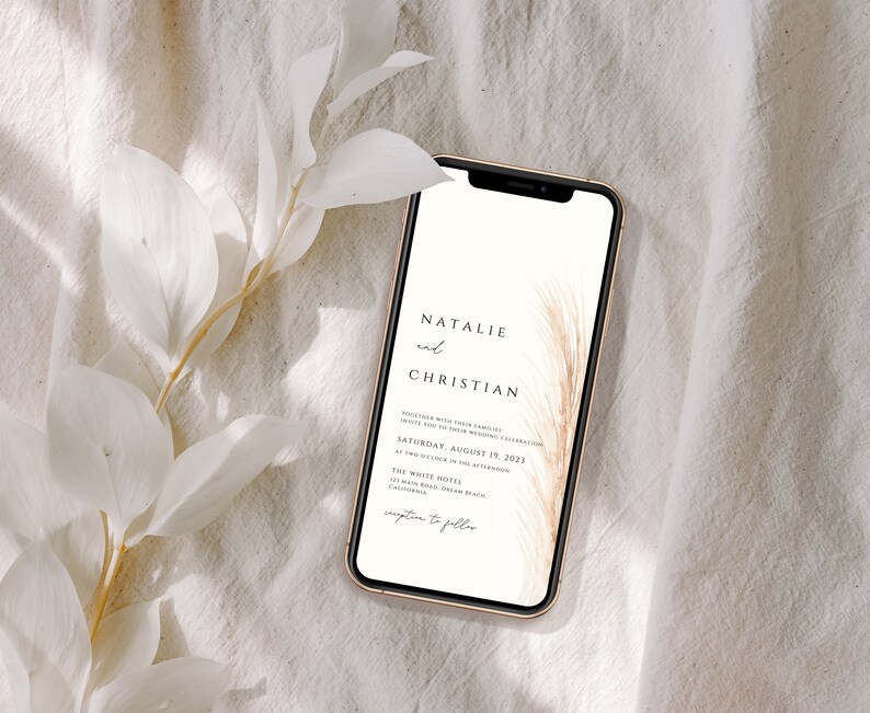 May include: A smartphone displays a wedding invitation with the names "Natalie and Christian" in elegant script. The invitation includes the date, time, and location, with a decorative feather design. The phone is set against a neutral, textured background with white foliage.