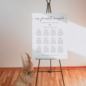 May include: A white seating chart with the text "our favorite people" and "PLEASE FIND YOUR SEAT" at the top. Below the text is a heart symbol and the names "OLIVIA & WILLIAM". The chart lists the names of guests assigned to each table number, from Table 1 to Table 12. The date "JUNE 25, 2022" and location "Temecula, CA" are printed at the bottom of the chart.