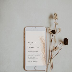 May include: A rose gold smartphone displays a wedding invitation with the names "Natalie and Christian" and event details. Dried floral elements are arranged beside the phone, creating a minimalist, elegant aesthetic.