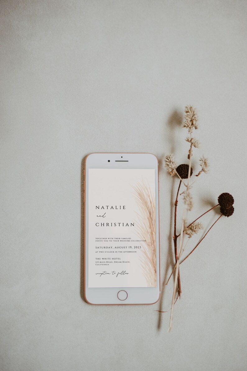 May include: A rose gold smartphone displays a wedding invitation with the names "Natalie and Christian" and event details. Dried floral elements are arranged beside the phone, creating a minimalist, elegant aesthetic.