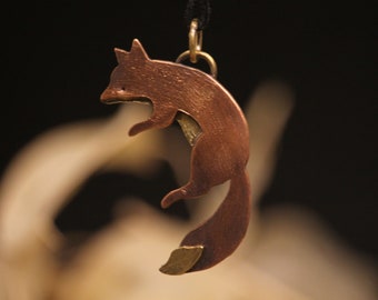 May include: A copper-colored fox pendant suspended from a black cord. The fox is depicted in a leaping pose, with a detailed silhouette. The pendant is attached to a gold-colored bail. The background is dark, highlighting the pendant.