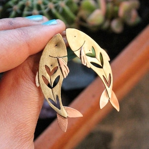 Op de afbeelding: Een paar oorbellen in de vorm van vissen in messingkleur. Elke oorbel heeft een uitgesneden bladerontwerp en koperkleurige vinnen. De oorbellen worden tegen een vage achtergrond van planten gehouden.