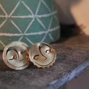 Peut inclure: Deux boucles d'oreilles dorées représentant le soleil et la lune. Les boucles d'oreilles sont circulaires, avec une surface texturée et des traits du visage découpés. La boucle d'oreille soleil a un bord festonné. Présentées sur une surface sombre.