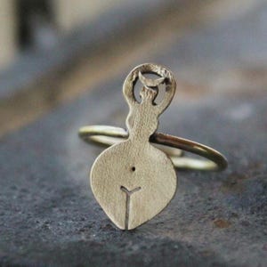 Peut inclure: Une bague de couleur laiton avec un design unique. La bague présente une figure stylisée avec un corps arrondi et une tête en forme de croissant. Le corps de la bague est fin et circulaire, et la figure est centrée.