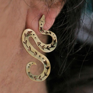 Può includere: Un orecchino color ottone a forma di serpente, con un design curvo. L'orecchino a serpente presenta ritagli a forma di foglia e piccoli dettagli a punti. L'orecchino è indossato sull'orecchio, con la testa del serpente rivolta verso l'alto.