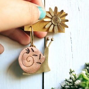Puede incluir: Pendientes hechos a mano con diseño de caracol y flor. El pendiente de caracol tiene una concha color cobre con detalles de hojas, mientras que el pendiente de flor es dorado. Se muestran sobre un fondo claro.