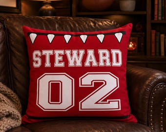 May include: A square red throw pillow featuring a white graphic of a pennant banner at the top, the name STEWARD in white block letters, and the number 02 in large white block digits with a black outline, centered on the front of the pillow.