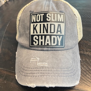 Puede incluir: Gorra de camionero gris desgastada con parte trasera de malla y visera curva. La parte delantera muestra el texto "NOT SLIM KINDA SHADY" en blanco y negro. La gorra tiene un aspecto vintage y desgastado.