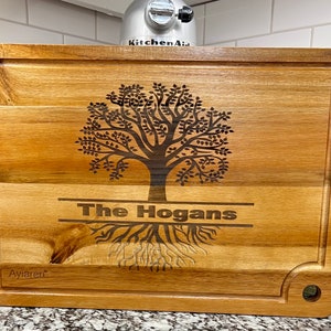 Può includere: Un tagliere in legno con un disegno di albero e il testo "The Hogans" inciso su di esso. Il tagliere ha un foro nell'angolo in alto a destra e il testo "Ayiaren" nell'angolo in basso a sinistra.