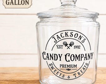 May include: A clear glass gallon jar with a lid featuring a black circular graphic that reads Jackson's Candy Company Est 1992 Premium Sweets & Treats with a small lollipop icon in the centre. The jar is transparent with a wide mouth and base.