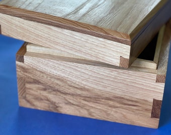 May include: A wooden box with a lid, made of light wood with a darker wood trim. The box is open and the lid is resting on top of the box.