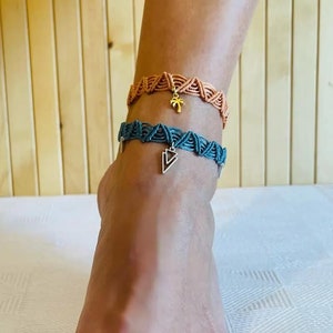 以下が含まれることがあります： チャーム付きのアンクレットが2つ。上のアンクレットは薄い茶色で、ヤシの木のチャームが付いています。下のアンクレットは濃い青色で、三角形のチャームが付いています。