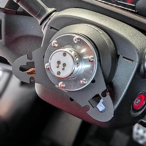 Op de afbeelding: Close-up van een zwarte stuurwielnaafadapter met een zilveren midden en meerdere bouten. De adapter is bevestigd aan een zwarte stuurkolom. De afbeelding toont een deel van het dashboard.