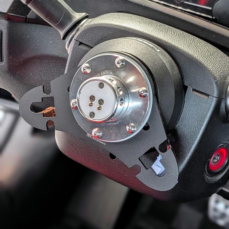 Op de afbeelding: Close-up van een zwarte stuurwielnaafadapter met een zilveren midden en meerdere bouten. De adapter is bevestigd aan een zwarte stuurkolom. De afbeelding toont een deel van het dashboard.