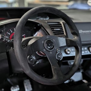 Op de afbeelding: Een zwart stuurwiel met een su&egrave;de-achtige grip is gemonteerd in het interieur van een voertuig. Het wiel heeft een cirkelvormig ontwerp met een centrale naaf en verschillende bedieningsknoppen. Het dashboard en andere interieuronderdelen zijn zichtbaar op de achtergrond.