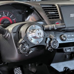 Op de afbeelding: Close-up van een aangepast autostuur met een zilveren en zwarte naaf en extra bedieningsmodules. De naaf is met bouten aan de stuurkolom bevestigd. Het dashboard en de meters zijn zichtbaar op de achtergrond.