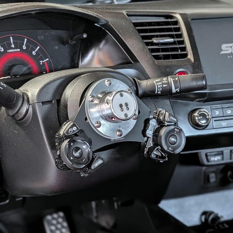 Op de afbeelding: Close-up van een aangepast autostuur met een zilveren en zwarte naaf en extra bedieningsmodules. De naaf is met bouten aan de stuurkolom bevestigd. Het dashboard en de meters zijn zichtbaar op de achtergrond.