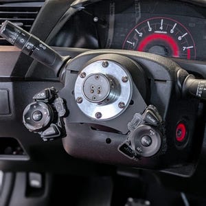 Op de afbeelding: Close-up van een autostuur met een aangepaste naaf. Het stuur is zwart met zilveren accenten, waaronder een cirkelvormige plaat met bouten. Bedieningsknoppen zijn zichtbaar aan de linker- en rechterkant, en het dashboard is op de achtergrond.