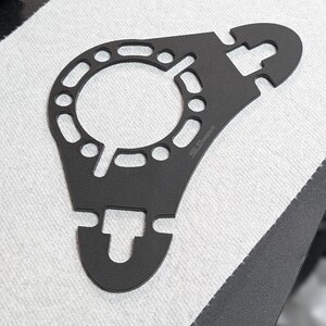 Op de afbeelding: Een zwart metalen gereedschap met een cirkelvormig midden en meerdere gaten. Het gereedschap heeft twee gebogen armen met open sleuven en de tekst "SLG Works" is zichtbaar. Het gereedschap is ontworpen voor een specifiek mechanisch doel.