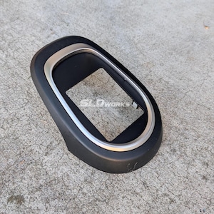 May include: A matte black plastic automotive trim bezel with a smooth, rounded oval shape and a metallic silver inner border. The center features a rectangular cutout, and the text SLDWORKS is printed in white on the inner black surface.