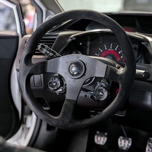 Op de afbeelding: Een zwart stuur met een su&egrave;de-achtige grip en een metalen middenstuk. Het stuur is gemonteerd in het interieur van een auto, met meters en bedieningselementen zichtbaar op de achtergrond. Het stuur heeft een sportief ontwerp.