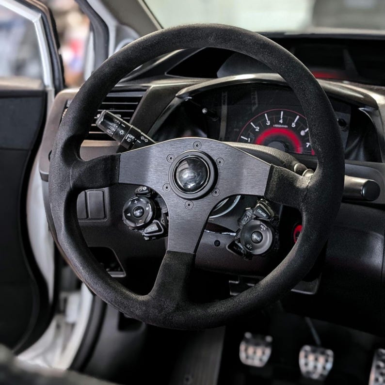 Op de afbeelding: Een zwart stuur met een su&egrave;de-achtige grip en een metalen middenstuk. Het stuur is gemonteerd in het interieur van een auto, met meters en bedieningselementen zichtbaar op de achtergrond. Het stuur heeft een sportief ontwerp.