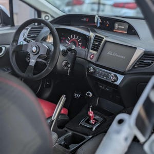 May include: The interior of a black car with a black steering wheel, a black dashboard, and a black gear shifter. The car has a red and black seat and a black center console. The dashboard has a digital display that says "SLD Works".