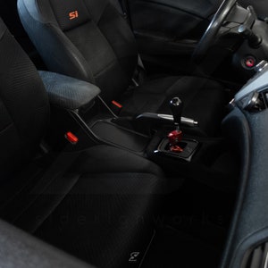 Peut inclure: Int&eacute;rieur noir d'une voiture avec un volant en cuir noir, des si&egrave;ges noirs et un levier de vitesses noir avec un pommeau rouge. Les si&egrave;ges ont un motif &agrave; carreaux noir et gris. La voiture a un embl&egrave;me "SI" rouge sur le si&egrave;ge passager.