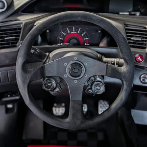 Op de afbeelding: Een zwart stuur met een su&egrave;de-achtige grip, een centrale naaf en bedieningsknoppen. Het dashboard achter het stuur toont een rode snelheidsmeter en andere meters. Het auto-interieur is donker, met diverse schakelaars en bedieningselementen zichtbaar.