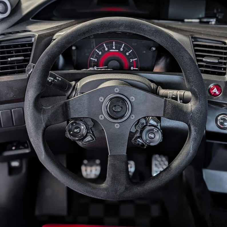 Op de afbeelding: Een zwart stuur met een su&egrave;de-achtige grip, een centrale naaf en bedieningsknoppen. Het dashboard achter het stuur toont een rode snelheidsmeter en andere meters. Het auto-interieur is donker, met diverse schakelaars en bedieningselementen zichtbaar.