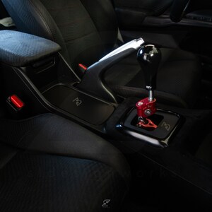 Peut inclure: Gros plan sur l'int&eacute;rieur d'une voiture en cuir noir avec un levier de vitesses argent&eacute; et noir. Le levier de vitesses a un levier rouge et un pommeau noir. L'int&eacute;rieur comprend &eacute;galement un accoudoir en cuir noir avec un bouton rouge et un panneau noir avec la lettre "R" dessus.