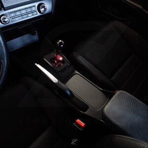 Peut inclure: Int&eacute;rieur de voiture noir avec un pommeau de levier de vitesse en cuir noir, un levier de vitesse argent&eacute; et une console centrale en cuir noir avec un bouton rouge.