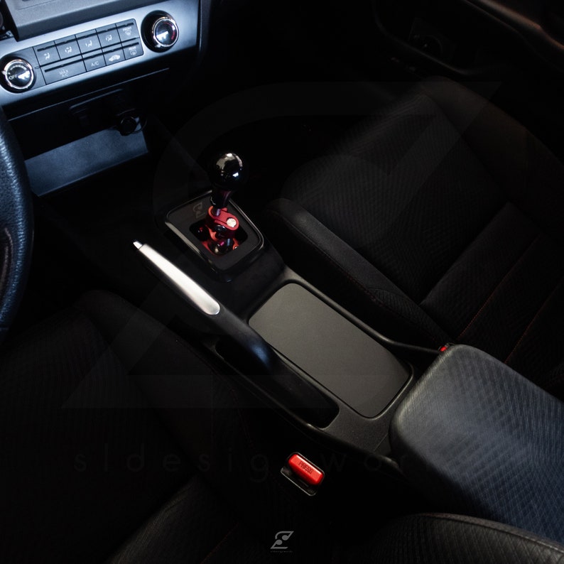 Peut inclure: Int&eacute;rieur de voiture noir avec un pommeau de levier de vitesse en cuir noir, un levier de vitesse argent&eacute; et une console centrale en cuir noir avec un bouton rouge.