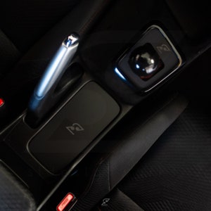 Peut inclure: Int&eacute;rieur de voiture en cuir noir avec un levier de vitesses argent&eacute;, un s&eacute;lecteur de vitesses noir et un compartiment de rangement noir avec un logo. Le compartiment a un bouton rouge marqu&eacute; "Press".