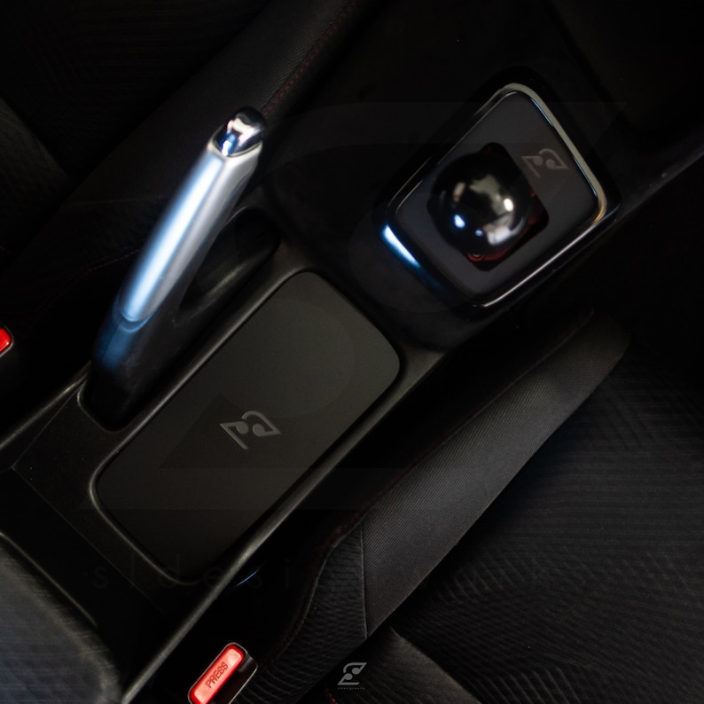 Peut inclure: Int&eacute;rieur de voiture en cuir noir avec un levier de vitesses argent&eacute;, un s&eacute;lecteur de vitesses noir et un compartiment de rangement noir avec un logo. Le compartiment a un bouton rouge marqu&eacute; "Press".