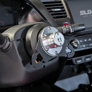 Op de afbeelding: Een zwart stuur met een zilveren en zwarte aftermarket stuurwieladapter ge&iuml;nstalleerd. De adapter heeft de tekst "SLD" en andere markeringen. Het dashboard en de bedieningselementen van de auto zijn zichtbaar op de achtergrond.