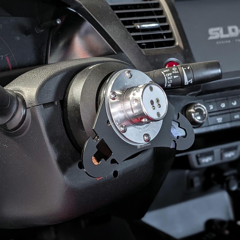 Op de afbeelding: Een zwart stuur met een zilveren en zwarte aftermarket stuurwieladapter ge&iuml;nstalleerd. De adapter heeft de tekst "SLD" en andere markeringen. Het dashboard en de bedieningselementen van de auto zijn zichtbaar op de achtergrond.