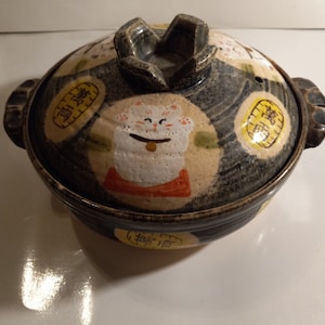 May include: A brown ceramic pot with a lid featuring a white cat with black eyes and a pink nose. The pot is decorated with yellow and black circles with Japanese characters.