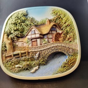 May include: A ceramic wall plaque depicting a cottage with a thatched roof, a water wheel, and a stone bridge over a stream. The scene is surrounded by lush greenery and a blue sky.