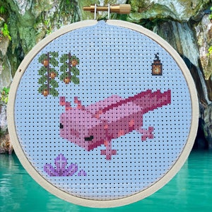 May include: Cross stitch pattern of a pink axolotl swimming in a blue pond with a green plant and a lantern. The pattern is in a wooden embroidery hoop. The image is in front of a cave and a body of water.