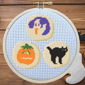 May include: A white embroidery hoop with a blue background and three Halloween-themed cross-stitch patterns: a ghost, a pumpkin, and a black cat. The patterns are surrounded by a white grid.