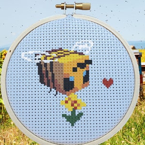 Könnte beinhalten: Kreuzstichmuster einer pixelartigen Biene mit einem Herzen auf blauem Hintergrund. Die Biene hält eine gelbe Blume. Das Muster ist in einem Reifen und ist von einem Feld mit Sonnenblumen und Bienenstöcken umgeben. Der Text "PDF" befindet sich in der unteren rechten Ecke des Bildes.