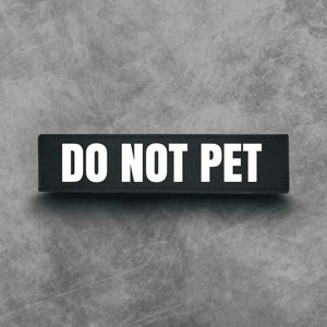 May include: A rectangular black patch with the white text "DO NOT PET" centered on it. The patch is set against a textured gray background.