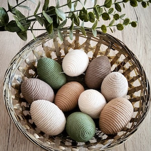 Könnte beinhalten: Ein Weidenkorb gefüllt mit gehäkelten Ostereiern in verschiedenen Brauntönen, Grün- und Cremetönen. Die Eier sind kreisförmig angeordnet, wobei die grünen Eier oben und unten im Korb liegen.