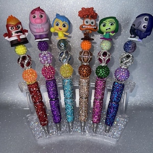 May include: Six pens featuring character toppers from the movie Inside Out. Each pen has a barrel covered in colorful rhinestones and is decorated with various textured, glittery, and metallic beads in matching color schemes.
