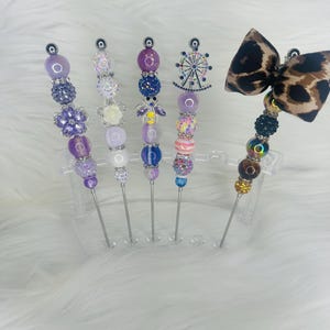 Puede incluir: Una colección de palillos de cóctel decorativos con diseños de cuentas. Los palillos presentan varios colores, incluyendo morado, blanco, rosa y marrón, con adornos como una noria, una abeja y un lazo con estampado de leopardo. Se exhiben en un soporte de plástico transparente.